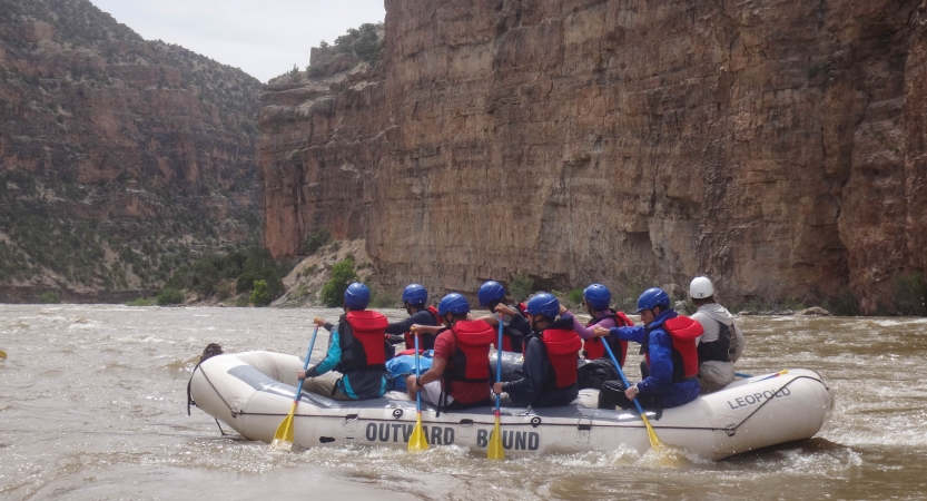 A group of people wearing safety gear paddle a raft. The river is framed by tall canyon walls. 